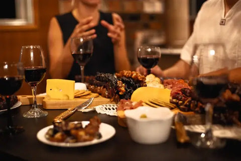 A photo of a couple friends sitting around a charcuterie board with churchkhela on it. they are also drinking wine and eating cheese with crackers and meats.