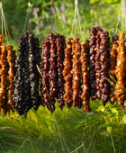Bundles of Churchkhela that are hung up on a lengthwise stick. The flavours from left to right go, apricot, grape, pomegranate, apple pie, pomegranate then apricot. The background is a grassy plane with some purple flowers in the back.