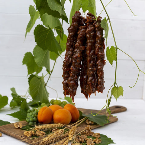 A full bundle of Churchkhela being hung up overtop of a wooden board with apricots, walnuts, grapes and wheat stocks on top of it. There are leaves in the background.
