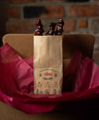 A brown bag with 3 Pomegranate Churchkhela inside. The bag is inside a shipping box that has burgundy gift wrap paper inside.