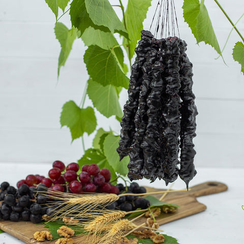 A full bundle of Churchkhela being hung up overtop of a wooden board with grapes, walnuts and wheat stocks on top of it. There are leaves in the background.