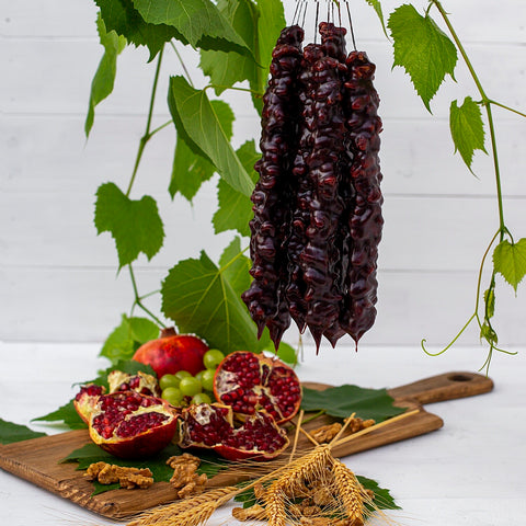 A bundle of pomegranate churchkhela being hung up white a wooden board underneath has pomegranates, walnuts and wheat stocks on it. There are leaves hanging in the background